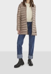 Vadderad knälång kappa i ljus taupe med dragkedja och vertikal quiltad design. Kombinerad med uppvikta denim och svarta ankelboots.