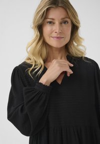 Juoda suknelė su tekstūruota, audinių struktūra. Turi V formos iškirptę, pūstas rankoves su mygtukų detalėmis ir susiraukšlėjusią, plazdančią sijoną.