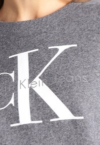 Sweatshirt gris avec un tissu texturé, arborant un grand "K" blanc et le texte "Klein Jeans" en dessous dans une police subtile.
