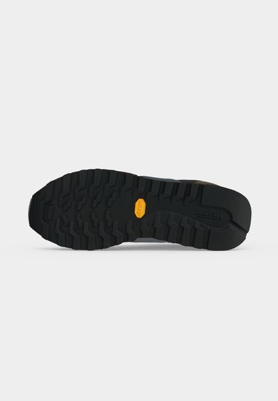 Semelle de chaussure noire robuste avec des crampons profonds et un logo Vibram jaune au centre sur un fond clair uni.