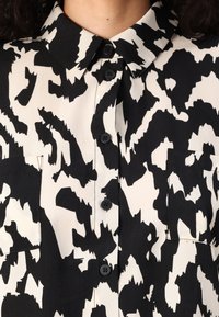 Chemise à motifs noir et blanc avec un col, comportant deux poches avant et des boutons noirs le long de la patte de boutonnage. Texture de tissu lisse.