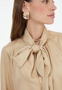 Beige blouse with a tied bow detail at the collar. Smooth, textured fabric, featuring long sleeves and a structured fit.