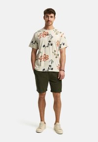 Chemise à manches courtes de couleur crème avec un imprimé floral orange, bleu et vert. Associée à un short vert foncé et des baskets claires.
