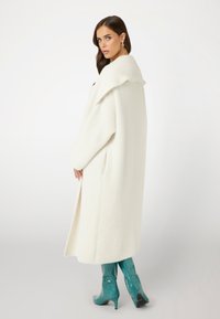Lange witte jas met een wolachtige textuur, voorzien van een grote kraag en zijzakken. Gecombineerd met glanzende teal, kniehoge, krokodillenpatroon laarzen.