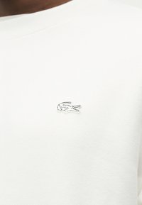 Chemise blanche avec un petit logo de crocodile brodé sur la poitrine gauche, portée par une personne à la peau foncée.