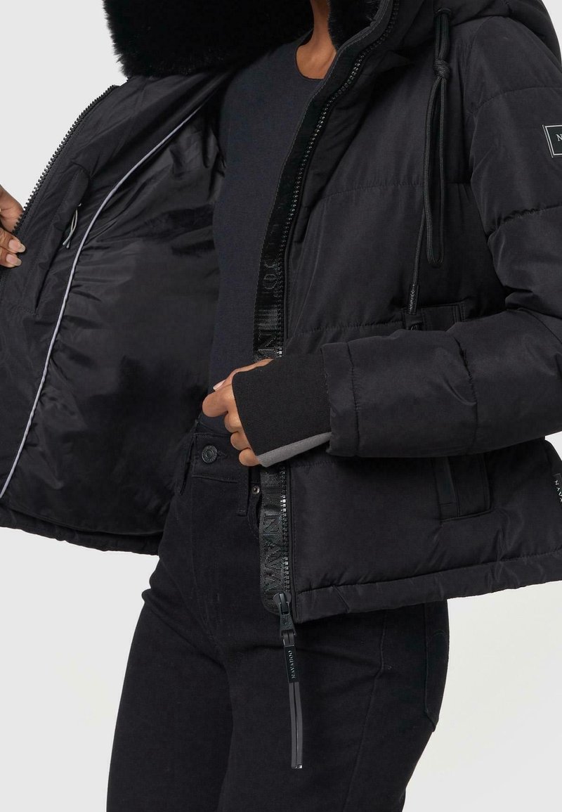 Veste matelassée noire avec capuche doublée de fourrure, zips et poignet intérieur gris contrastant. Dispose d'une ouverture frontale à glissière et de poches latérales.