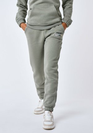 Un pantalon de survêtement vert clair en tissu doux, doté de poignets élastiques, de poches latérales et d'un accent logo sur la cuisse gauche. Stylé avec des baskets blanches.