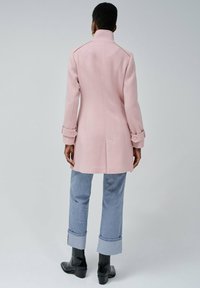 Abrigo de lana rosa claro con corte recto, puños abotonados y cuello alto, combinado con pantalones de mezclilla remangados y botas negras hasta el tobillo.
