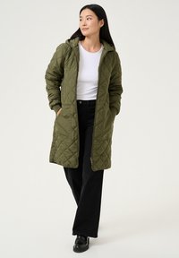 Parka matelassée vert olive avec capuche, manches longues, poches latérales et fermeture éclair intégrale à l'avant. Portée sur un haut blanc et un pantalon noir.