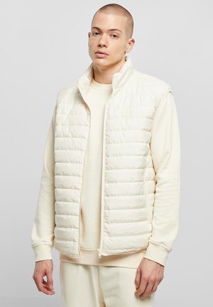 Vesta zateplená krémovej farby s prešívaným dizajnom, vysokým golierom a zapínaním na zips. Kombinovaná so zodpovedajúcou mikinou s okrúhlym výstrihom.