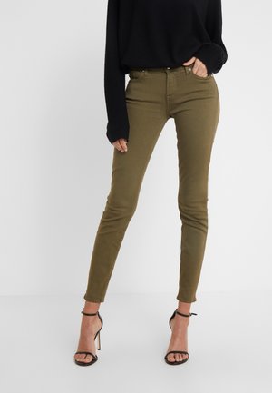 Vrouw draagt olijfgroene strakke jeans met een zwart top met lange mouwen en zwarte sandalen met hoge hakken, staand tegen een effen achtergrond.