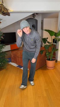 Homme portant un pull zippé gris, un pantalon vert foncé et des chaussures argentées, posant dans un salon avec une plante et un canapé.