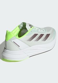 Scarpa da corsa leggera con una tomaia in rete verde menta, strisce riflettenti marroni e un accento sul tallone di un verde brillante. Presenta una suola intermedia bianca e una suola testurizzata.