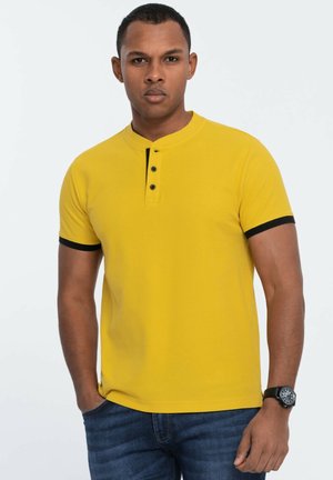 Geel polo shirt met korte mouwen, zwarte accenten en een drie-knopen sluiting. Getextureerde stof, relaxte pasvorm, gecombineerd met donkerblauwe jeans.