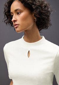Top bianco con scollo alto e apertura a forma di chiave. Texture morbida, maniche corte, design minimale e senza motivi visibili.