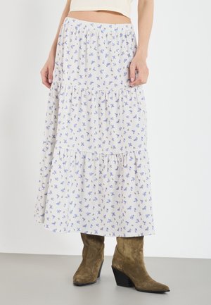 Femme portant une jupe midi blanche à volants avec un petit imprimé floral bleu et des bottes de cowboy en daim marron, cropped top pas entièrement visible.