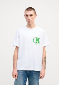 Camiseta de algodón blanca con mangas cortas, que presenta un logo verde de CK en el lado izquierdo del pecho. Se lleva con jeans azules.