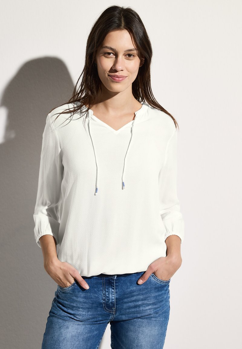 Lichte witte blouse met een V-hals, een trekkoordcapuchon en driekwart mouwen, gecombineerd met blauwe denim jeans. Soepele stoftextuur.