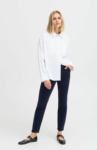 Camicia bianca con bottoni e due tasche sul petto, abbinata a pantaloni slim-fit blu navy. Il modello indossa scarpe nere piatte. Design semplice e pulito.