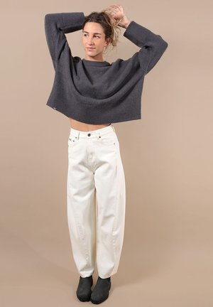 Femme portant un pull court gris foncé, un pantalon blanc taille haute et des bottines noires, ajustant ses cheveux devant un fond beige.