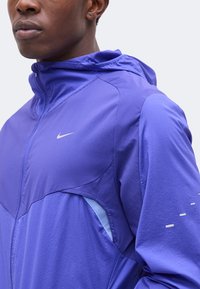 Gros plan d'une personne portant une veste Nike violette zippée avec capuche, montrant une ventilation latérale doublée de mesh et des détails réfléchissants sur la manche.