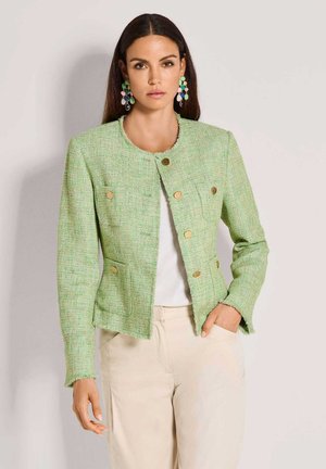 Vrouw draagt een pastelgroene gestructureerde jas met gouden knopen, witte top, beige broek en kleurrijke hangende oorbellen, staand tegen een effen achtergrond.