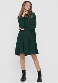 Robe verte à col avec des manches longues, un bas à volants et une coupe décontractée. Portée avec des bottes de combat à lacets noires. Texture de tissu lisse.