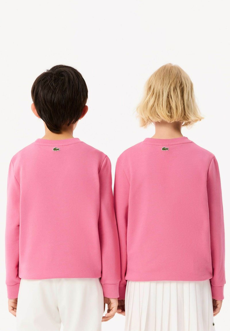 Rosa Sweatshirts mit langen Ärmeln, Rundhalsausschnitt, gerippten Bündchen und Lacoste-Logo auf der Rückseite, kombiniert mit weißen Hosen.