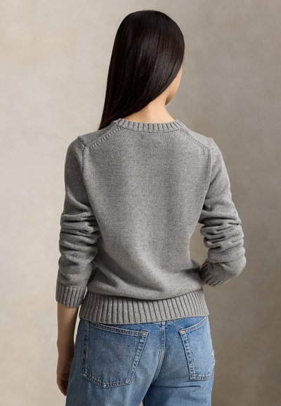 Femme aux longs cheveux foncés portant un pull en laine gris et un jean bleu, de dos sur un fond neutre.