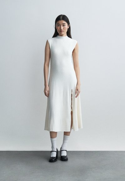 Femme debout contre un mur gris, portant une robe midi blanche sans manches à côtes avec une fente latérale, des chaussettes blanches et des chaussures Mary Jane noires.