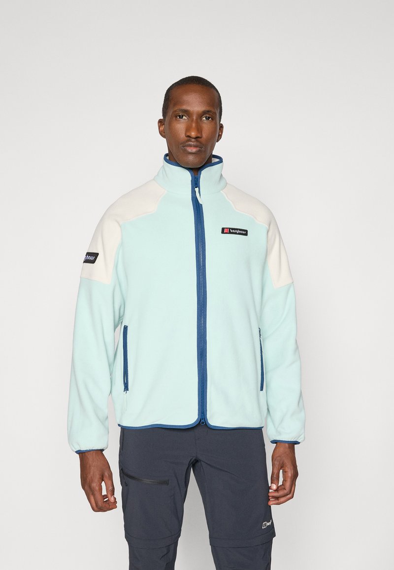 Man draagt een lichtblauwe en witte jas met rits en twee met rits afsluitbare zakken, en donkere broek, staand tegen een effen achtergrond.