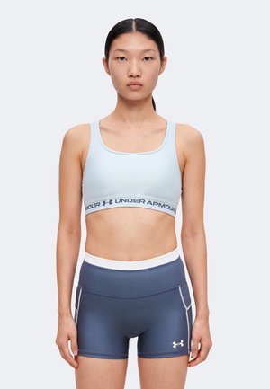 Ung kvinde, der vender fremad, iført en lyseblå Under Armour sports-bh og marineblå stramme atletikshorts med hvid taljebånd og logo.