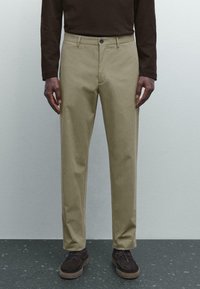 Man som bär beige raka byxor, mörkbrun långärmad skjorta och mörkbruna mockasneakers stående på ett fläckigt golv.