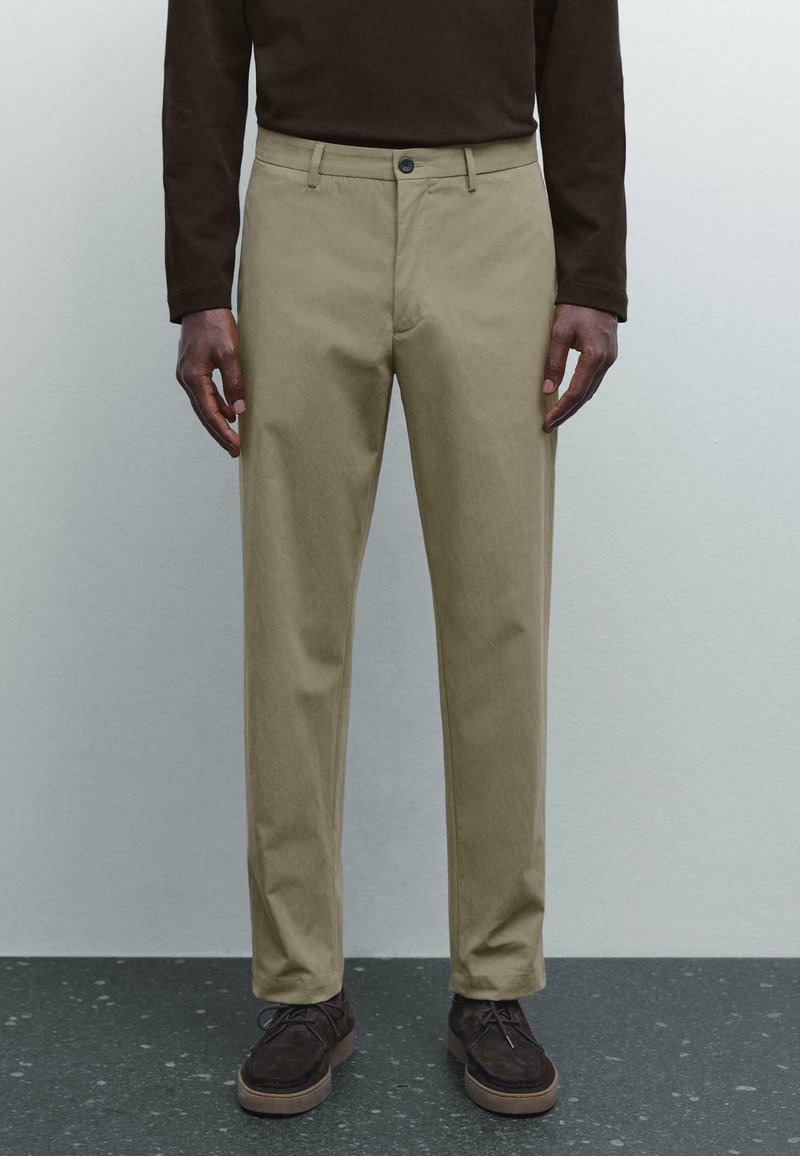 Man som bär beige raka byxor, mörkbrun långärmad skjorta och mörkbruna mockasneakers stående på ett fläckigt golv.