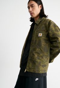Veste camouflage vert olive avec fermeture éclair, deux poches latérales et un petit patch logo ; portée sur une chemise noire et un pantalon sombre.