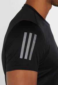 Czarna koszulka sportowa z krótkim rękawem o teksturowanym wykończeniu, z trzema refleksyjnymi szarymi paskami na lewym ramieniu.