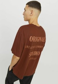 Camiseta de algodón marrón con texto blanco en la parte posterior: "ORIGINALS", "CREATIVE COMMUNITY" y "STUDIO". Manga corta y corte relajado.