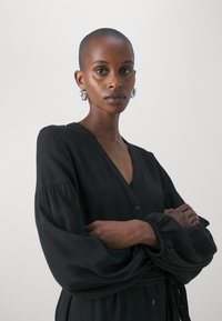 Black, long-sleeve button-up dress with a deep V-neck, gathered cuffs, and a tied waist; smooth texture and minimalist design.
