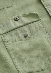 Light green fabric with a textured surface, featuring a pocket with a snap button closure and detailed stitching.