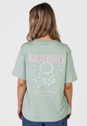 Kvinde set bagfra iført en lysegrøn T-shirt med lyserød tekst "VACACIONES" og havtema-tegninger inklusive en muslingeskal og en søstjerne.