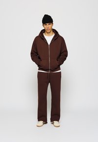 Brauner Zip-Hoodie und passende Jogginghose aus Baumwollmischung mit elastischen Bündchen und lockerer Passform, kombiniert mit beigen Turnschuhen.