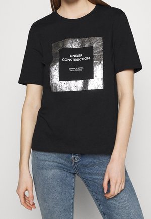 Osoba ubrana w czarną koszulkę z krótkim rękawem z srebrnym kwadratem i napisem "UNDER CONSTRUCTION BEWARE, A BETTER ME IS COMING", w połączeniu z niebieskimi dżinsami.