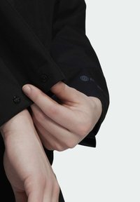 Des mains ajustant le poignet d'une chemise noire à manches longues avec des boutons visibles et un petit logo au niveau du poignet.