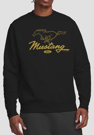 Mann trägt schwarzen Pullover mit goldfarbenem Ford Mustang-Logo, das ein galoppierendes Pferd und geschwungenen Text auf der Brust zeigt.