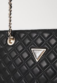 Bolso de mano negro acolchado con una brillante cadena dorada y una insignia de logo triangular. Presenta una superficie texturizada y costuras en forma de diamante.