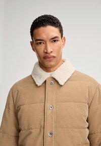 Giacca in corduroy color sabbia con collare in pelliccia sintetica color crema, dotata di chiusura con bottoni e design trapuntato. Texture liscia e forma strutturata.