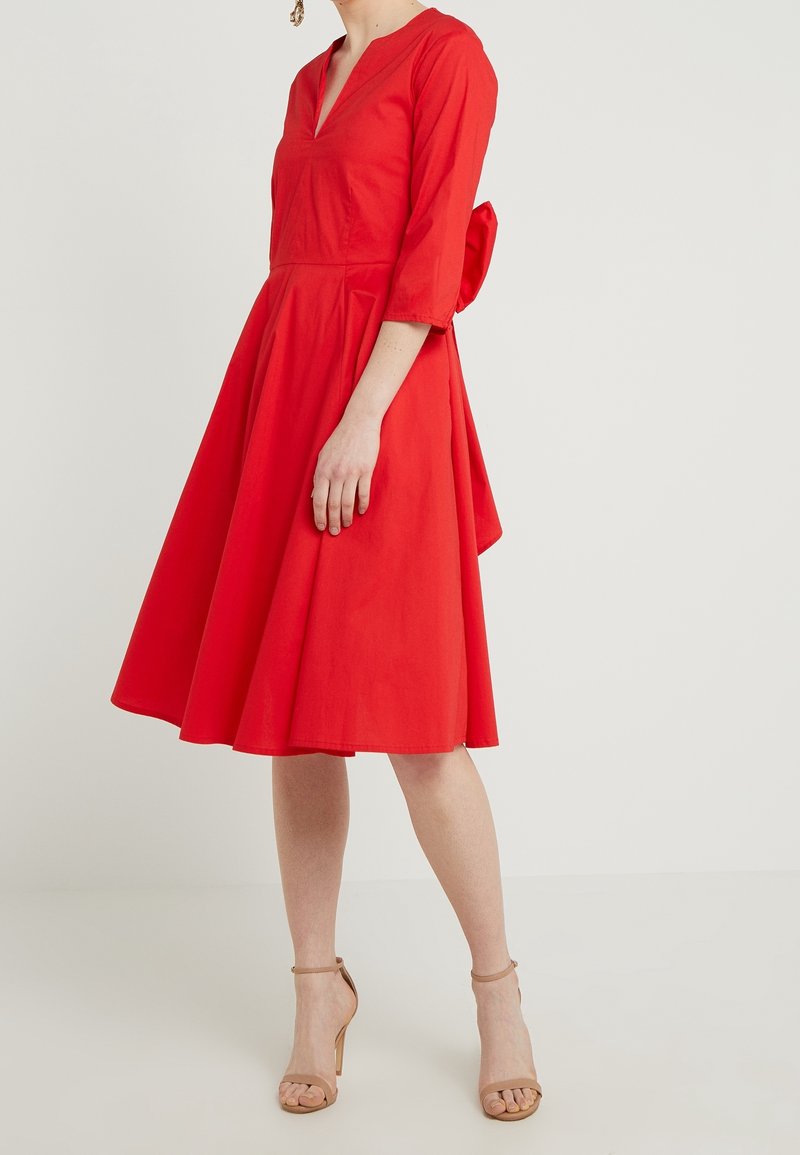 Femme portant une robe rouge longueur genou avec des manches trois-quarts et des sandales à talons hauts couleur nude, debout devant un fond uni.