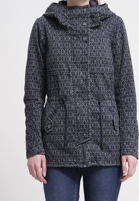 Marinblå jacka med mönster, huva, dragsko i midjan och dubbla knappar. Har en geometrisk design och framfickor. Strukturerad textil.