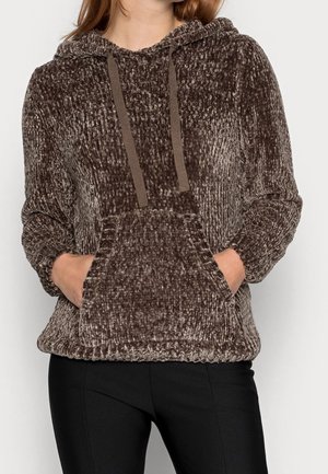 Kvinde iført en brun tekstureret chenille-hoodie med frontlomme og snøre, kombineret med sorte bukser, stående mod en ensartet baggrund.