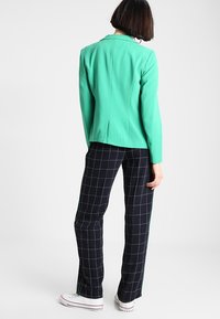 Blazer vert au design structuré, assorti à un pantalon noir à carreaux. La veste présente un col cranté et une silhouette ajustée.